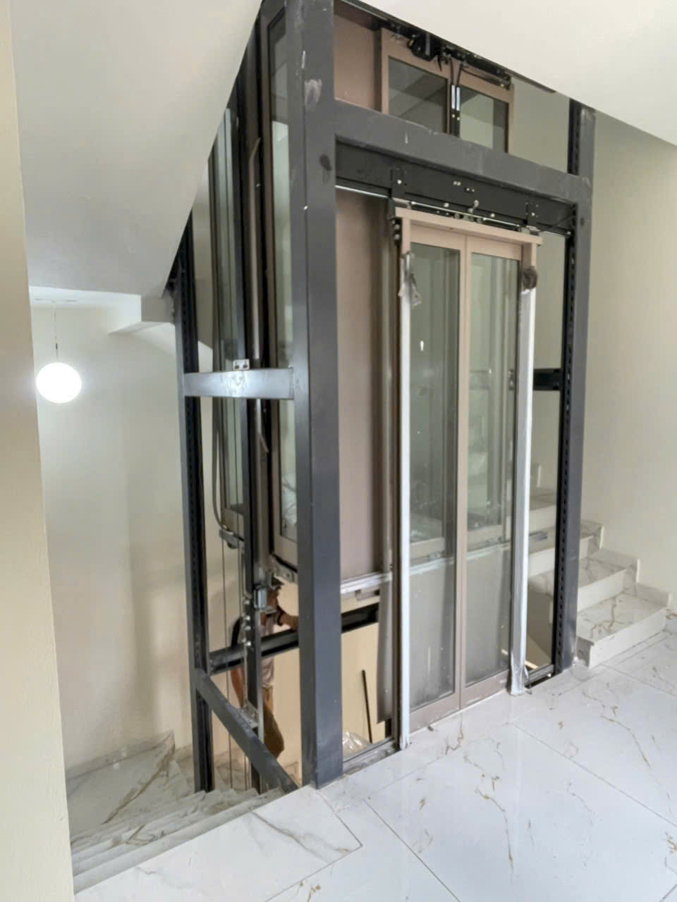 Thang máy gia đình khung thép đang lắp đặt giữa cầu thang trong nhà sàn đá marble